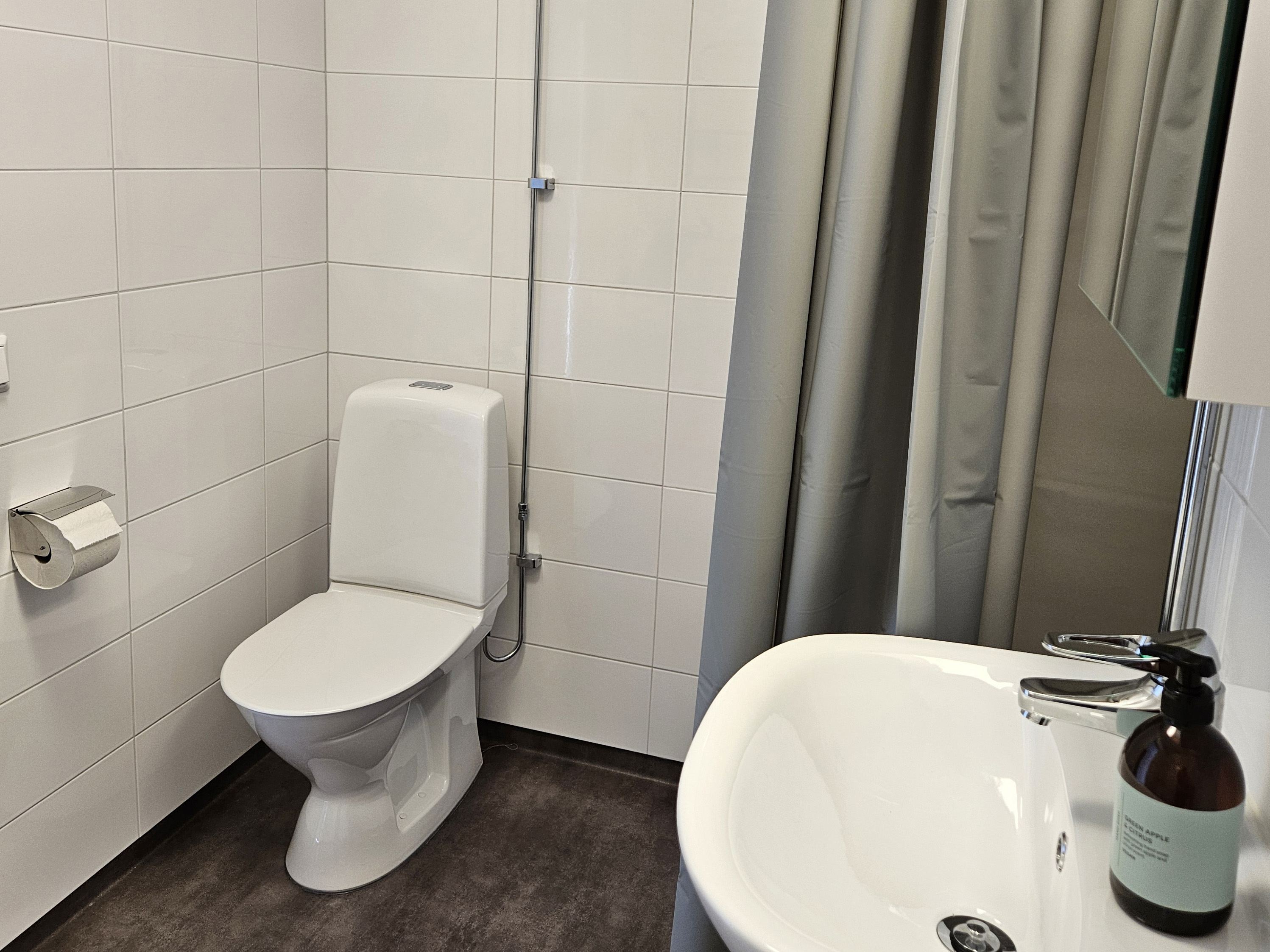 Bild på helrenoverat badrum på Sellerigången, väggar med stora vita kakelplattor, mörkt melerat plastgolvgolv. Grått duschdraperi.