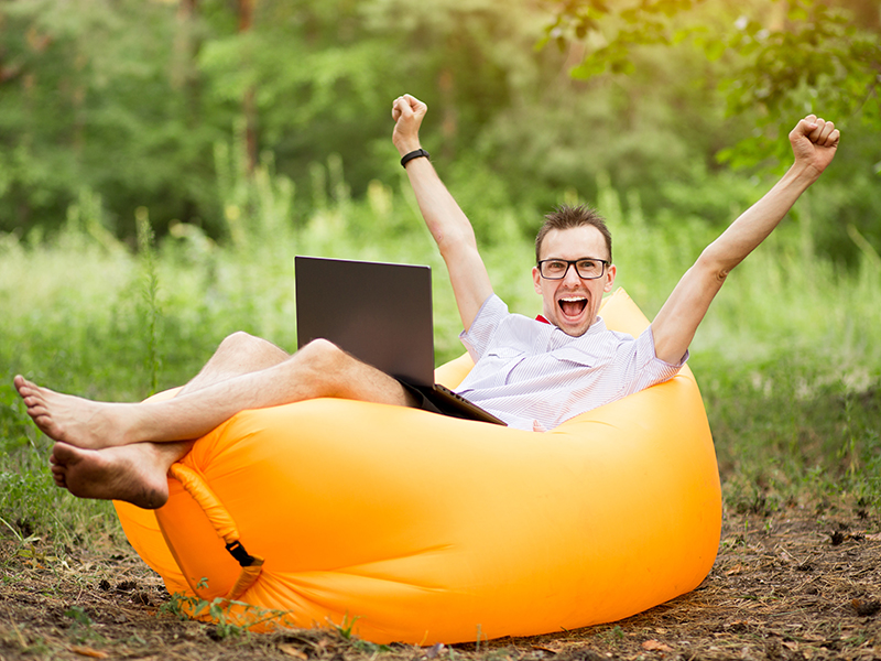 Lycklig man som sitter utomhus på en uppblåst säck med laptop i knät.